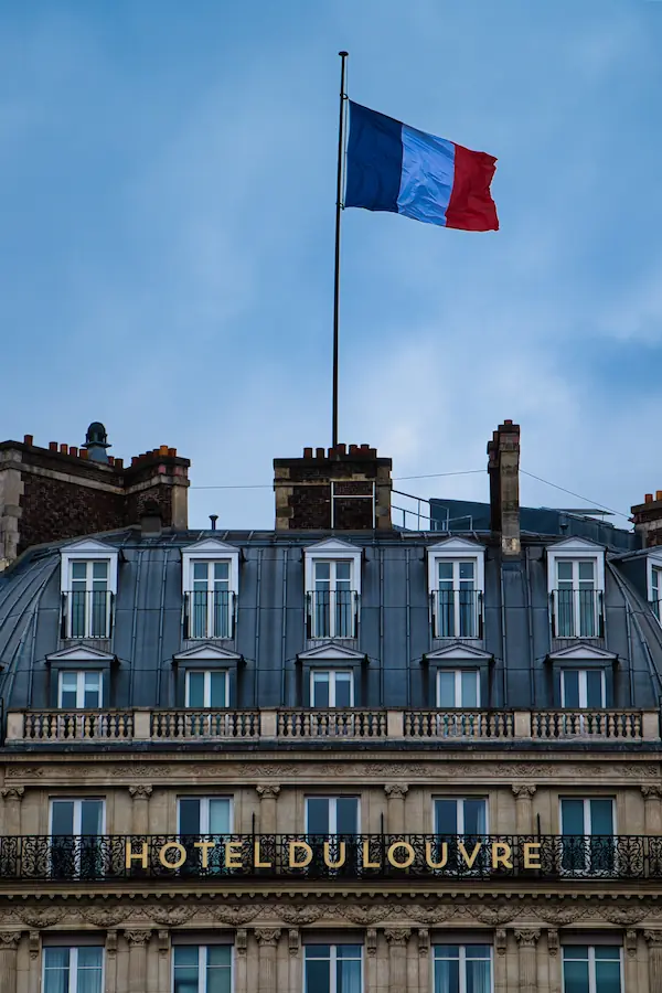 Façade haussmannienne de l'Hôtel du Louvre avec son toit mansardé en zinc, ses fenêtres alignées et le drapeau français flottant au sommet sous un ciel bleu parisien