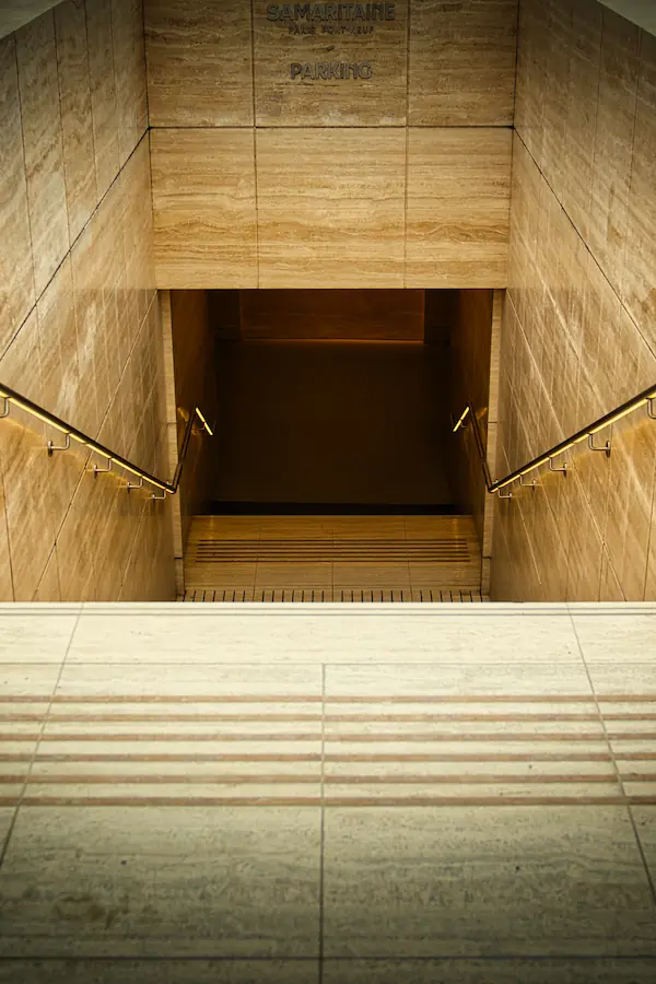 Escalier descendant vers le parking Samaritaine, habillé de marbre beige aux tons chauds, avec mains courantes métalliques dorées et éclairage tamisé