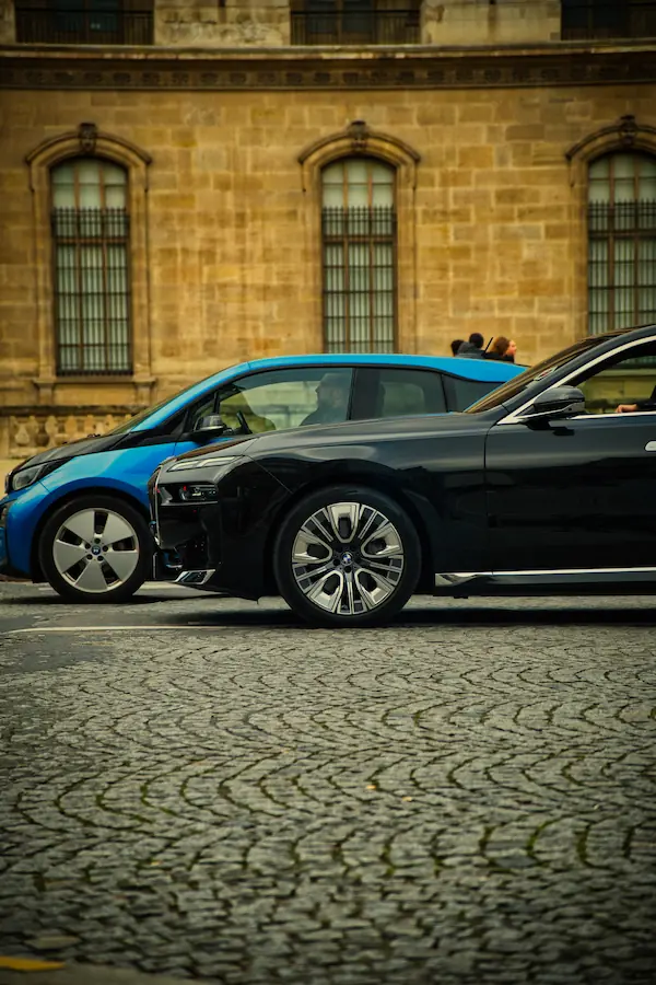 BMW électrique noire et BMW i3 bleue garées devant une façade historique en pierre, sur des pavés parisiens disposés en arêtes de poisson