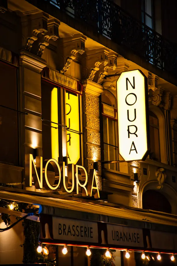 Enseigne lumineuse "NOURA" d'une brasserie libanaise, éclairée de nuit sur une façade haussmannienne ornementée, avec une guirlande de lumières en devanture