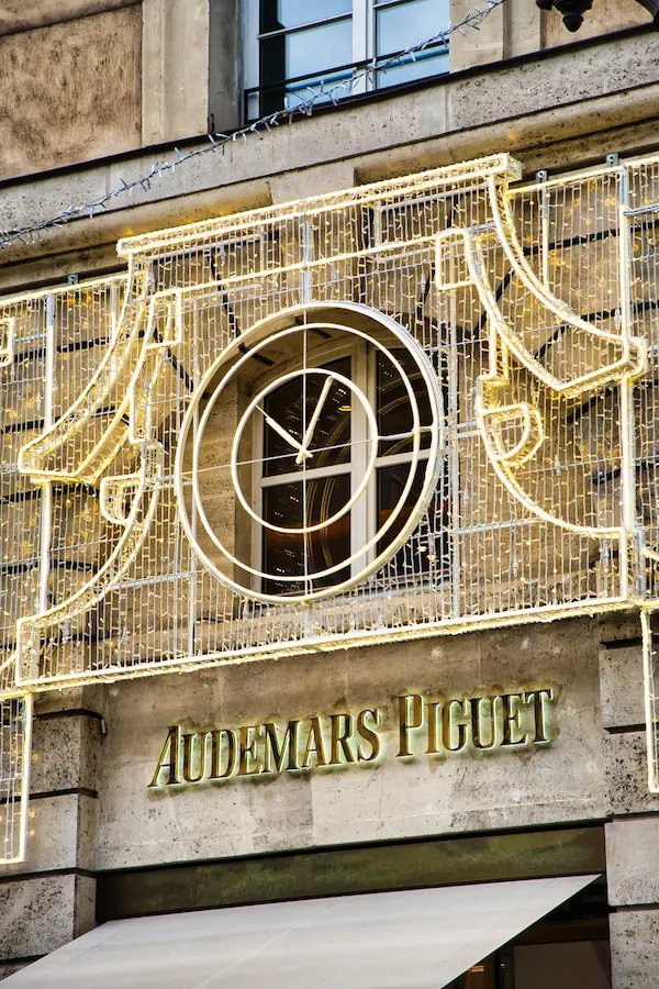 Façade de la boutique Audemars Piguet avec son enseigne dorée, surmontée d'une fenêtre circulaire en forme d'horloge et décorée de guirlandes lumineuses LED pour les fêtes