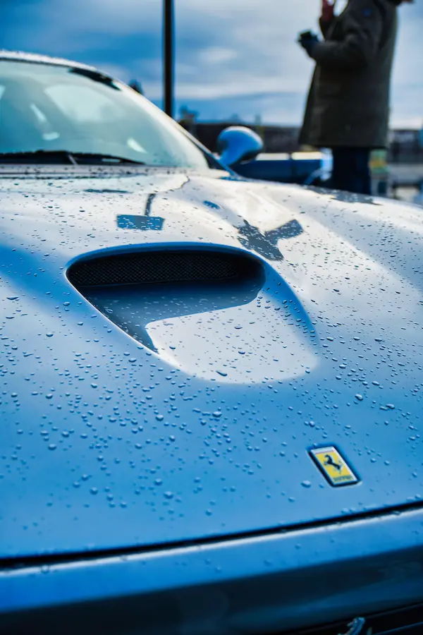 Détail du capot d'une Ferrari grise sous la pluie, montrant une prise d'air sculpturale et l'emblématique écusson du cheval cabré, avec des gouttes d'eau parsemant la carrosserie
