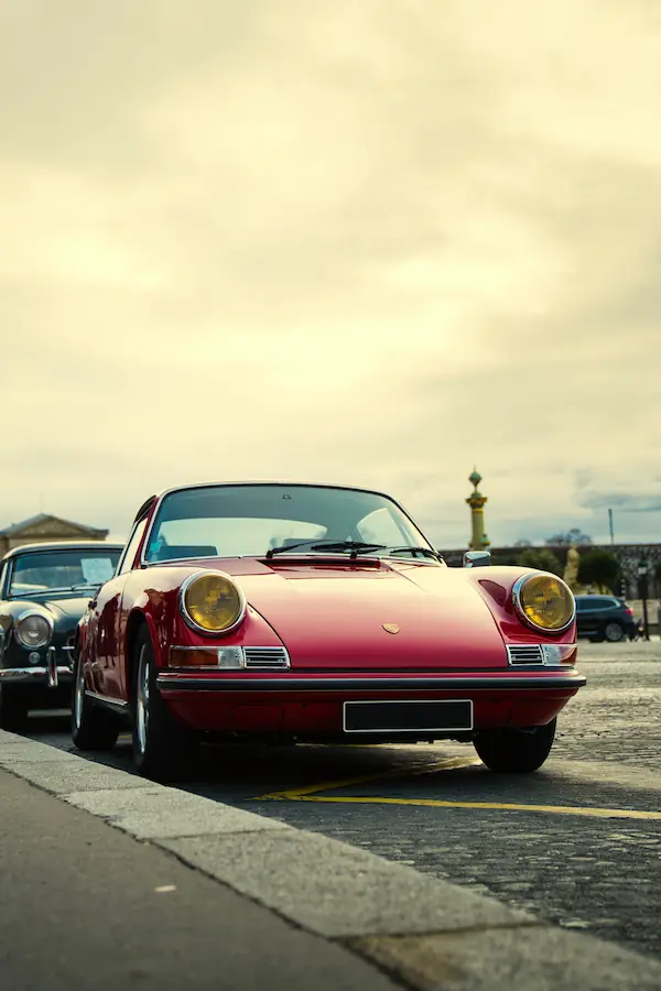 Porsche 911 classique rouge bordeaux avec phares jaunes, photographiée de face sur les pavés parisiens, incarnant l'élégance intemporelle du design automobile des années 60