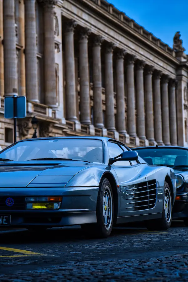 Ferrari Testarossa grise photographiée devant une façade néoclassique à colonnes, illustrant le contraste entre l'architecture classique parisienne et le design italien des années 80