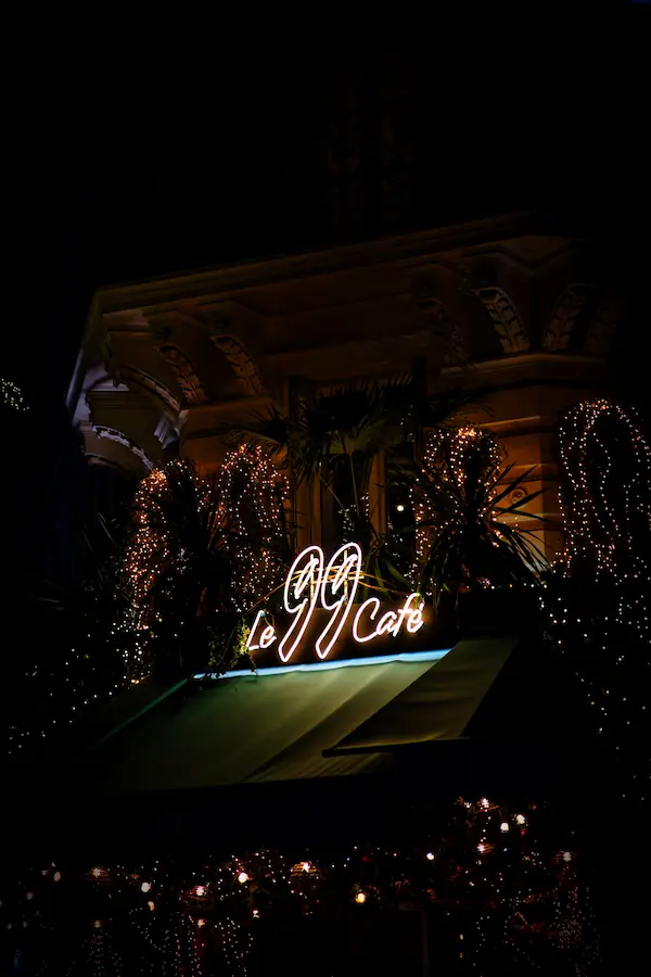 Enseigne lumineuse "Le 99 Café" en néon blanc, entourée de décorations de Noël scintillantes et de palmiers illuminés, créant une ambiance festive et chaleureuse dans la nuit parisienne