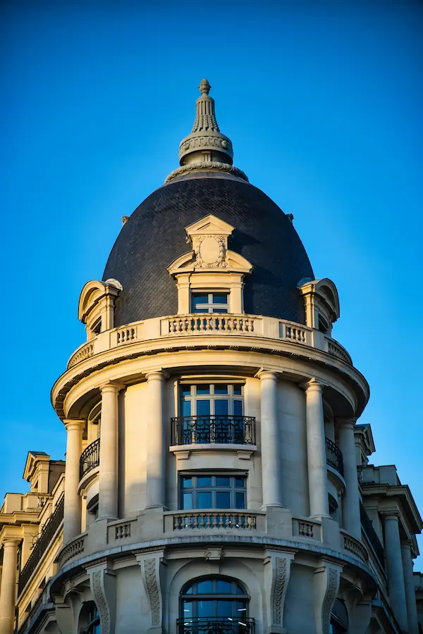 Dôme d'angle haussmannien avec son toit en ardoise noire et sa structure en pierre blanche, orné de colonnes, balcons en fer forgé et lucarnes sculptées, photographié sous une lumière dorée contre un ciel bleu intense