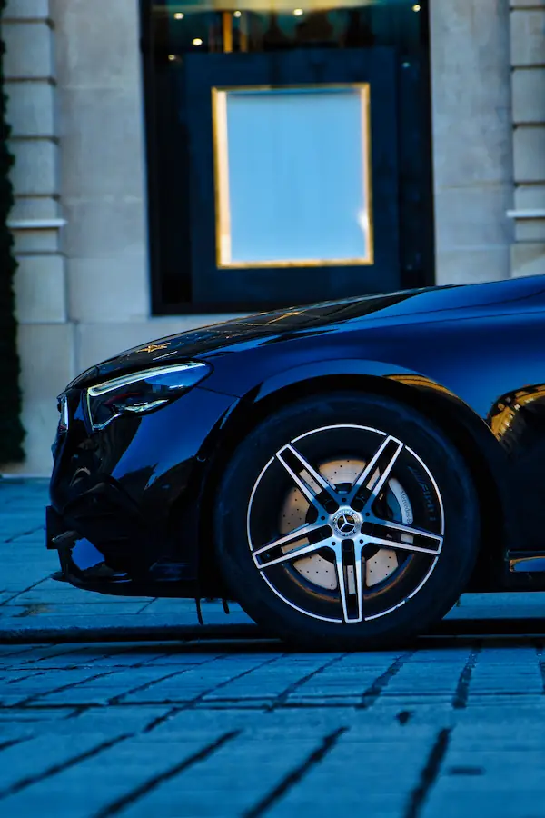 Détail d'une Mercedes-Benz noire montrant sa jante à cinq branches bicolore et son disque de frein perforé, photographiée devant une vitrine élégante au cadre doré