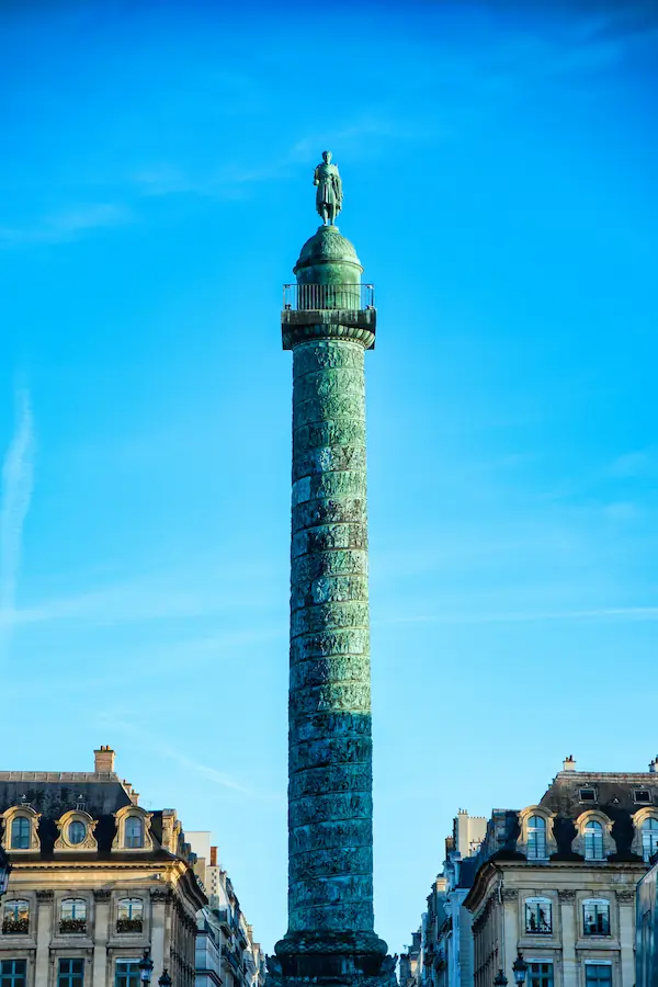 Colonne Vendôme à Paris, monument en bronze aux reliefs spiralés relatant les victoires napoléoniennes, surmontée d'une statue de Napoléon et encadrée d'immeubles haussmanniens sous un ciel bleu éclatant