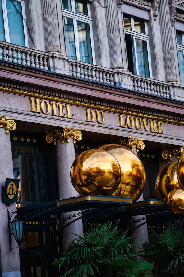 Entrée de l'Hôtel du Louvre avec son enseigne dorée, ses colonnes ornementées et ses caractéristiques sphères dorées réfléchissantes au-dessus de la marquise, encadrées de palmiers et de lanternes art déco