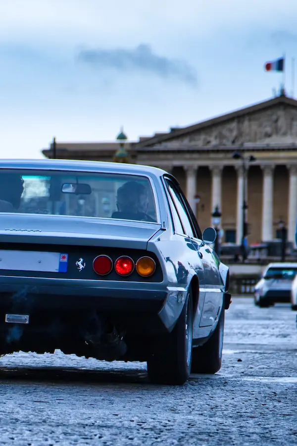 Arrière d'une Ferrari 365 GT4 2+2 grise photographiée devant une façade néoclassique parisienne, mettant en évidence ses feux arrière caractéristiques à trois optiques rondes et son emblème du cheval cabré