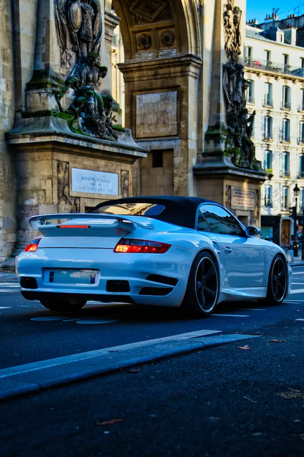 Porsche 911 Turbo blanche personnalisée, photographiée de trois-quarts arrière devant une façade historique parisienne ornée de sculptures, mettant en valeur son aileron caractéristique et ses jantes noires sportives