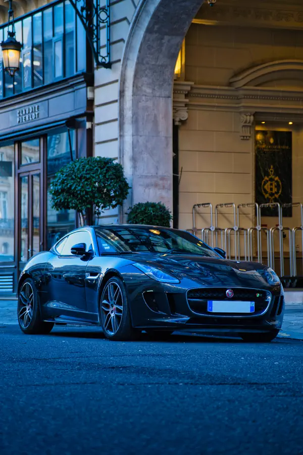 Jaguar F-Type coupé grise stationnée devant une façade haussmannienne parisienne, vue de trois-quarts avant, avec des arbustes en pot et une entrée voûtée en pierre en arrière-plan.
