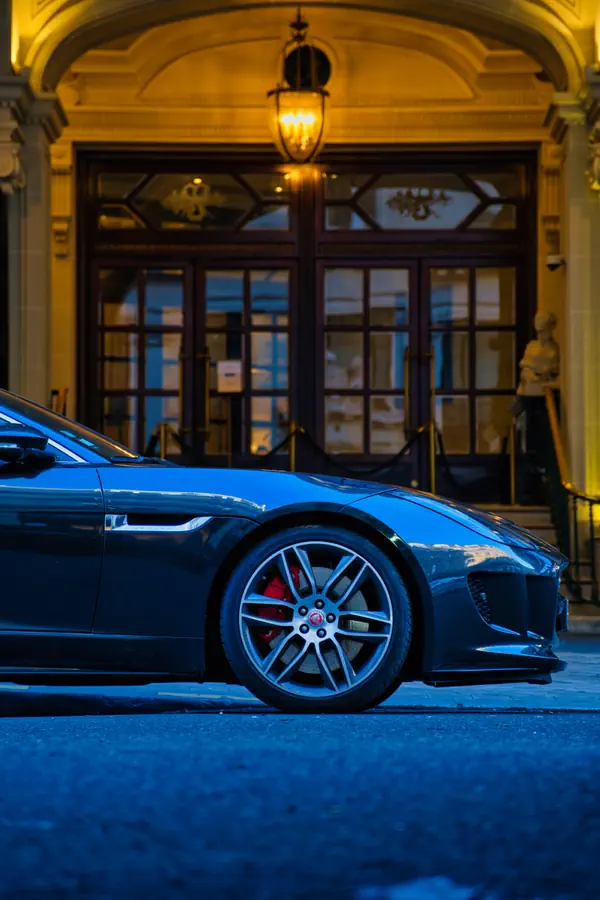Détail du profil avant d'une Jaguar F-Type grise, mettant en évidence la jante en alliage à cinq branches avec étrier de frein rouge, photographiée devant une entrée d'immeuble art déco éclairée par une lanterne classique.