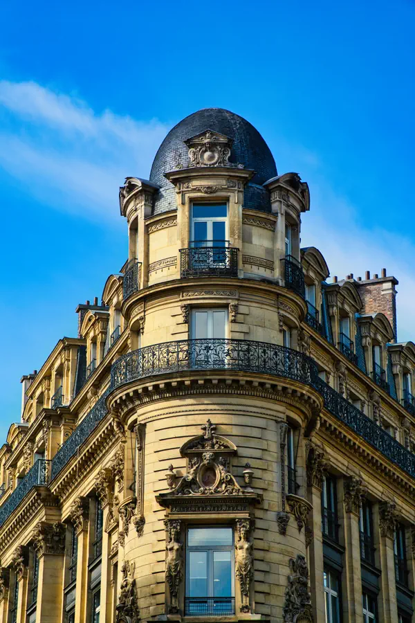Angle d'un immeuble haussmannien parisien avec son dôme d'ardoise caractéristique, orné de balcons en fer forgé, de cariatides et de sculptures décoratives, photographié en contre-plongée sous une lumière dorée et un ciel bleu vif.