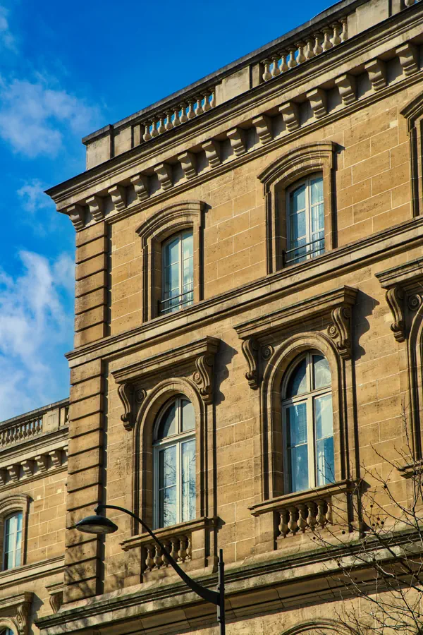 Façade d'un bâtiment historique en pierre de taille, style Second Empire, avec fenêtres cintrées ornées de consoles sculptées, balustrades décoratives et corniche à modillons, photographiée sous une lumière dorée et un ciel bleu.