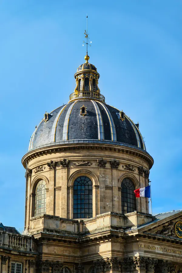 Dôme de l'Institut de France à Paris, avec son architecture classique en pierre de taille, sa coupole en ardoise ornée de bandes dorées, son lanternon à colonnes et sa girouette, surmonté du drapeau français, photographié sous un ciel bleu éclatant.