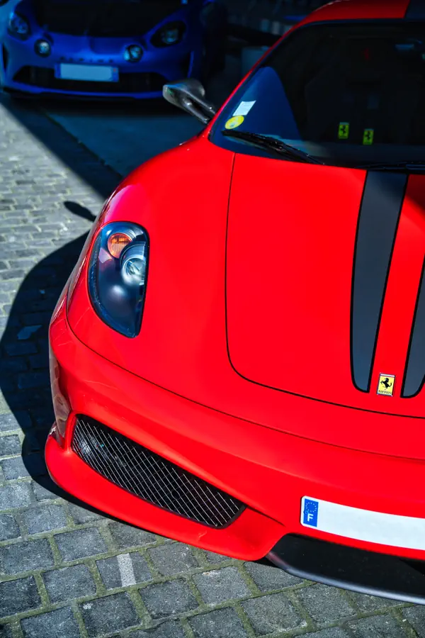 Détail avant d'une Ferrari F430 Scuderia rouge, montrant le phare caractéristique, la bande décorative noire, l'écusson Ferrari et la calandre inférieure en nid d'abeille, photographiée sur des pavés parisiens.
