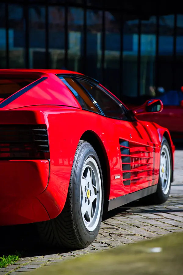 Vue latérale d'une Ferrari Testarossa rouge, mettant en évidence les caractéristiques prises d'air latérales à lamelles, la jante à cinq branches en alliage blanc et le profil emblématique de ce modèle des années 80.