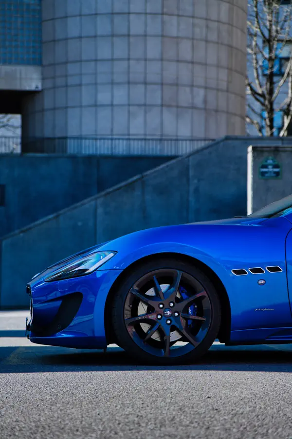 Partie avant d'une Maserati GranTurismo bleue avec jantes noires, stationnée devant un bâtiment moderne à façade quadrillée, montrant les trois ouïes caractéristiques sur l'aile.