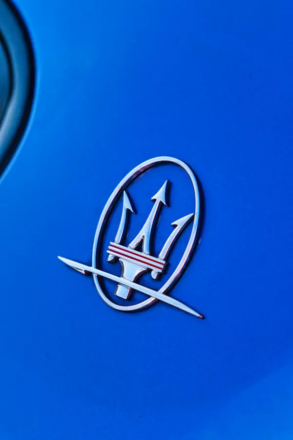 Logo Maserati chromé avec trident emblématique sur carrosserie bleue, montrant le détail du badge métallique avec ses trois flèches et bandes rouges caractéristiques.