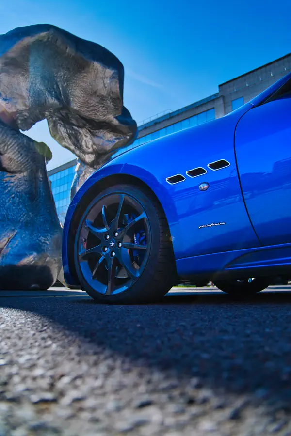 Maserati GranTurismo bleue avec jantes noires photographiée à ras du sol, montrant les trois ouïes latérales caractéristiques et l'inscription Pininfarina, à côté d'une sculpture métallique moderne devant un bâtiment contemporain.