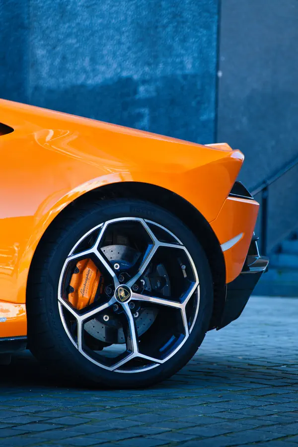 Détail de la roue arrière d'une Lamborghini orange avec jante en alliage au design géométrique, étrier de frein orange, disque de frein perforé et logo Lamborghini visible au centre de la jante, sur un sol pavé.