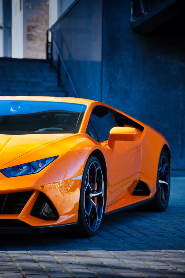 Lamborghini Huracán orange vif stationnée sur un pavage bleuté, photographiée de trois-quarts avant, montrant ses lignes agressives, phares angulaires et jantes géométriques, avec un mur sombre et un escalier en arrière-plan.