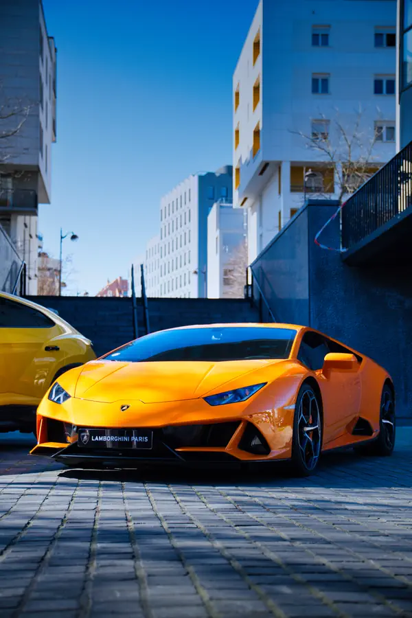 Lamborghini Huracán orange stationnée sur une place pavée dans un environnement urbain, avec plaque d'immatriculation Lamborghini Paris, immeubles modernes en arrière-plan et une partie d'une voiture jaune visible à gauche.