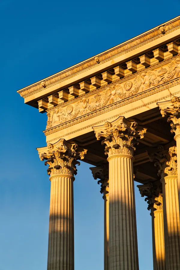 Détail architectural d'un temple néoclassique avec colonnes corinthiennes cannelées et entablement richement orné de frises sculptées, baigné dans une lumière dorée du soleil levant contre un ciel bleu profond.
