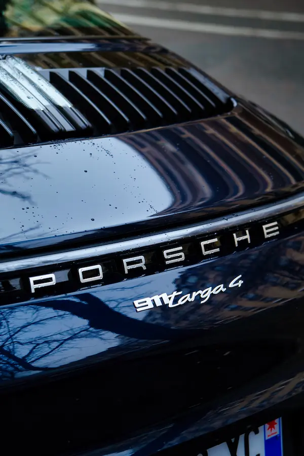 Détail de l'arrière d'une Porsche 911 Targa 4 noire, montrant les emblématiques lettres "PORSCHE" chromées et la grille de ventilation, avec des gouttes de pluie sur la carrosserie brillante
