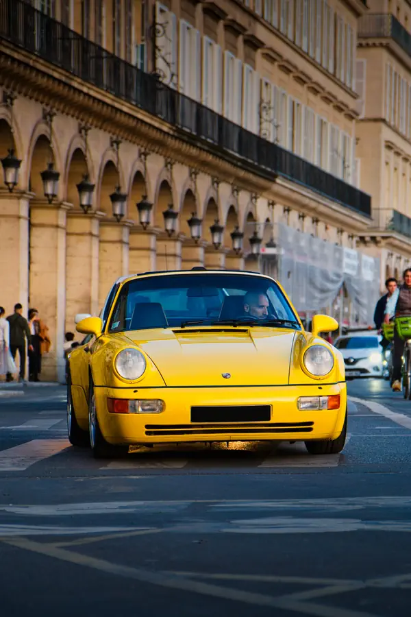 Porsche 911 jaune traversant un passage piéton dans une rue urbaine bordée d'architecture classique avec arcades et lanternes.