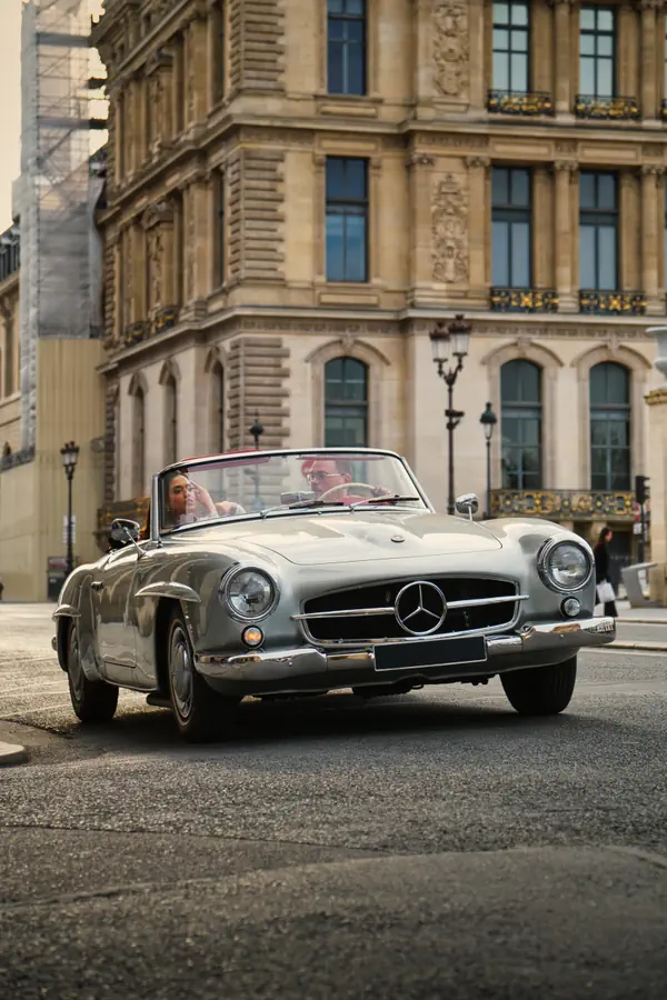 Mercedes-Benz 190 SL classique décapotable argentée circulant dans une rue parisienne, avec deux personnes à bord, devant un bâtiment historique orné de façade haussmannienne avec balcons en fer forgé et lampadaires d'époque.