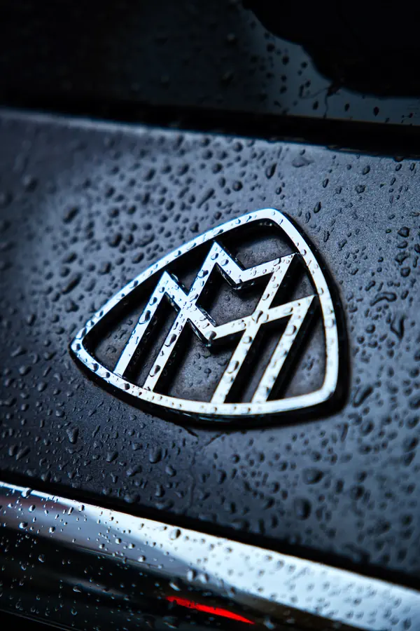 Gros plan sur le logo Maybach chromé sur une carrosserie noire couverte de gouttelettes d'eau, montrant l'emblème en forme de bouclier avec le double M stylisé caractéristique de la marque de luxe.