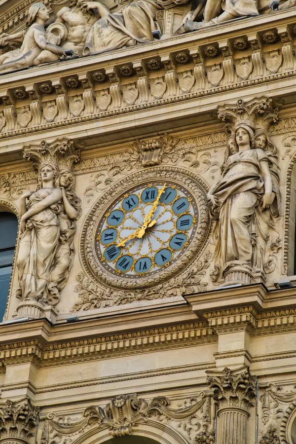 Horloge ornementale sur une façade de bâtiment historique parisien, avec cadran bleu et chiffres romains dorés, entourée de sculptures féminines allégoriques en pierre et de riches décorations architecturales en style Beaux-Arts.