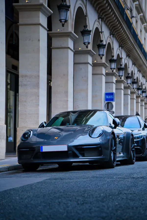 Porsche 911 gris foncé stationnée dans une rue parisienne élégante, devant une façade classique avec arcades et lanternes.