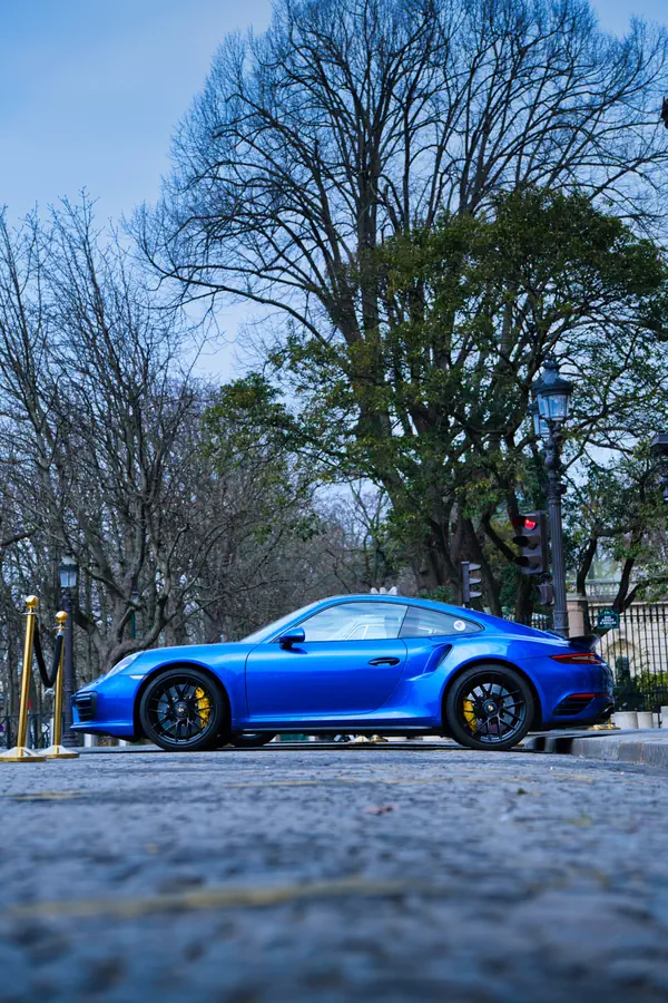 Porsche 911 Turbo bleue métallisée stationnée devant un parc parisien, avec jantes noires et étriers de frein jaunes, photographiée de profil avec des arbres en arrière-plan.
