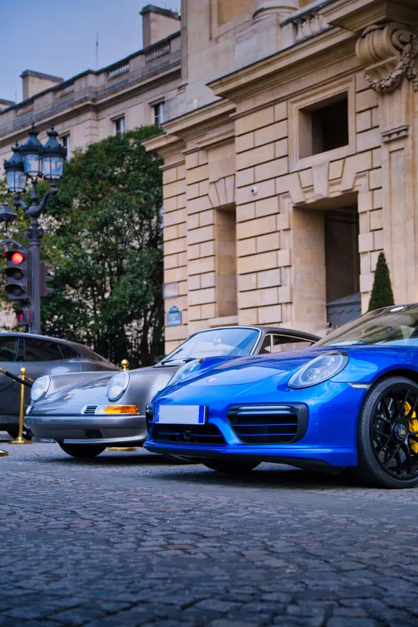 Deux générations de Porsche 911 stationnées côte à côte sur des pavés parisiens : un modèle classique gris argenté à l'avant et un modèle moderne bleu au premier plan.