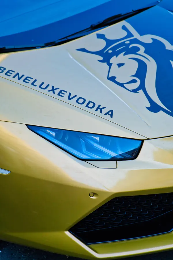 Gros plan sur l'avant d'une Lamborghini jaune personnalisée avec le logo bleu "BENELUXE VODKA" et un emblème de lion stylisé sur le capot, montrant le phare angulaire caractéristique et une partie de la calandre noire.