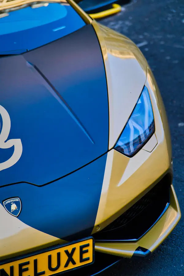 Gros plan sur le phare avant et l'emblème Lamborghini d'une Huracán personnalisée aux couleurs jaune et bleue, montrant les lignes angulaires caractéristiques du capot et la signature lumineuse LED.