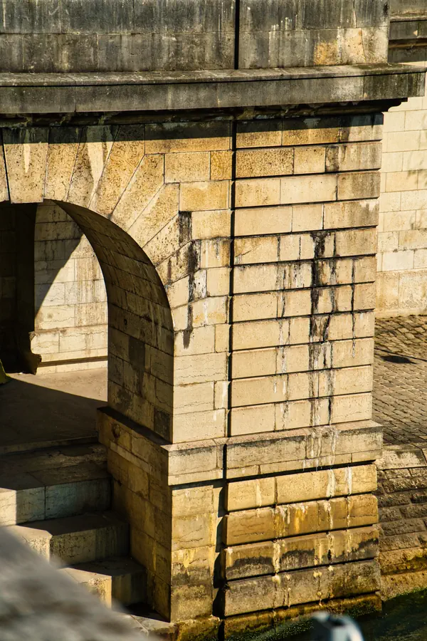 Détail architectural d'un pont parisien en pierre de taille, montrant une arche et un pilier massif avec des traces de patine et d'écoulement d'eau, illuminé par la lumière dorée du soleil, avec des pavés visibles à la base de la structure.
