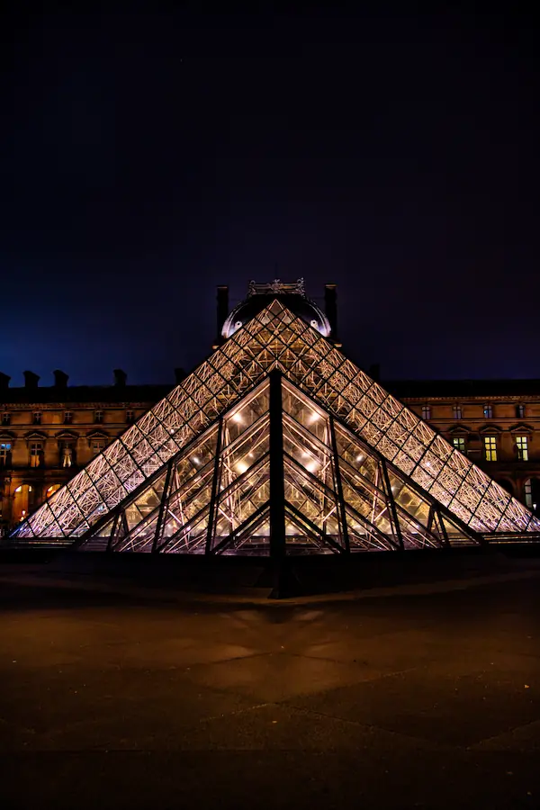 Pyramide du Louvre illuminée de nuit, sa structure géométrique en verre et métal brillant d'une lumière dorée, se détachant sur la façade historique du musée et un ciel nocturne profond