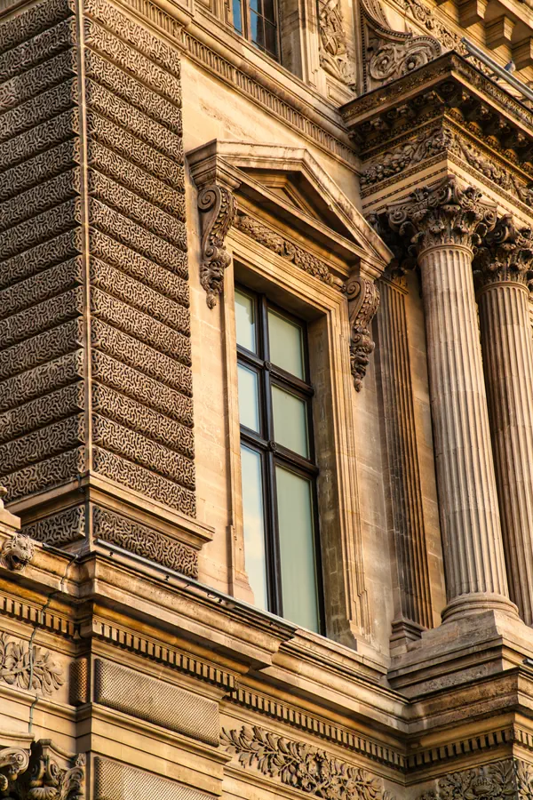 Détail architectural d'un bâtiment parisien historique avec façade ornementée, montrant un contraste entre une section à bossages décoratifs et une partie avec colonnes corinthiennes cannelées, fenêtre à fronton triangulaire et riches moulures sculptées, baigné dans une lumière dorée.