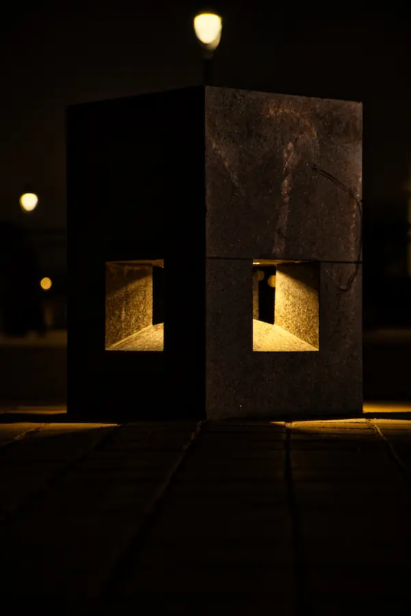 Borne lumineuse moderne en pierre noire photographiée de nuit, avec deux ouvertures rectangulaires éclairées créant un jeu d'ombre et de lumière géométrique sur le sol