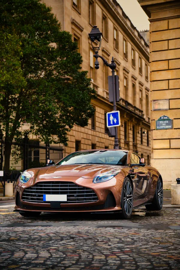 Aston Martin DB11 cuivre métallisé stationnée sur des pavés mouillés, avec façades haussmanniennes en pierre de taille.