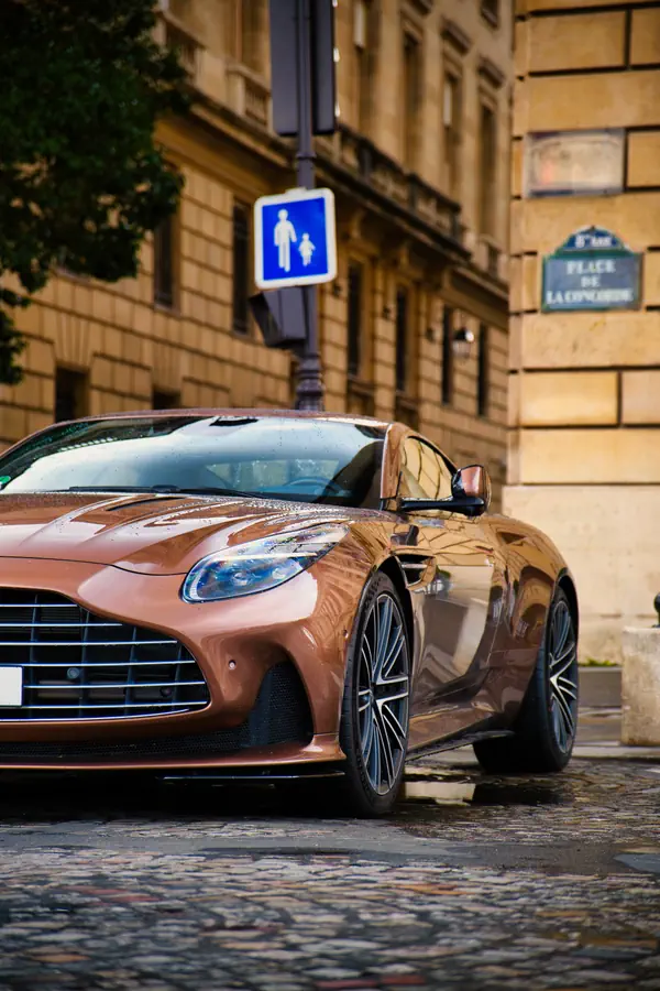 Aston Martin DB11 couleur bronze photographiée de trois-quarts avant sur les pavés humides.
