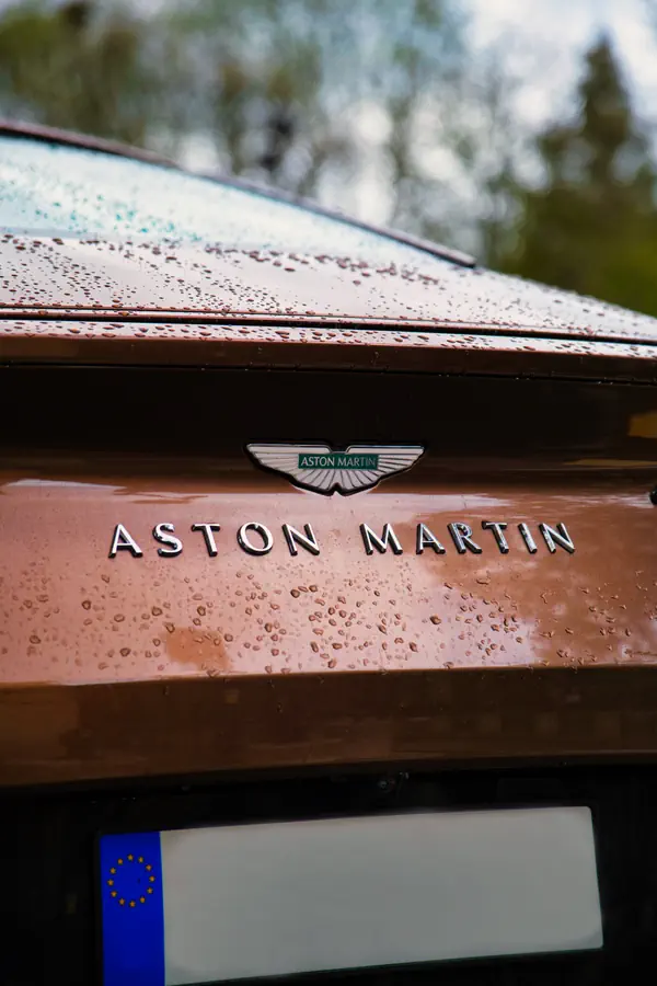 Gros plan sur l'arrière d'une Aston Martin bronze/cuivre montrant le logo ailé et le nom de la marque en lettres chromées, avec des gouttelettes d'eau sur la carrosserie.