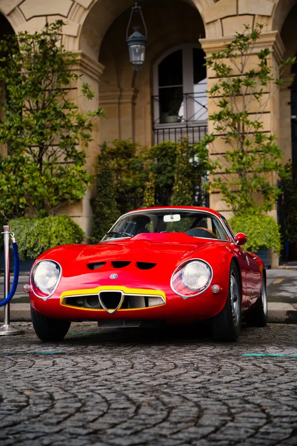Alfa Romeo Giulia TZ classique rouge vif stationnée sur des pavés devant un bâtiment historique avec arcades en pierre, entouré d'arbustes verts et de petits arbres, montrant sa silhouette emblématique avec phares ronds et calandre triangulaire.