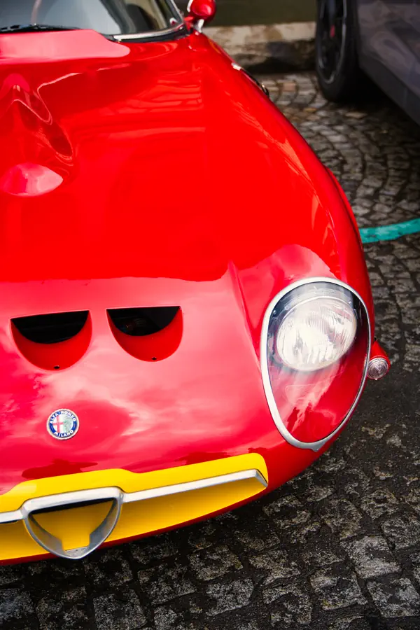 Gros plan sur l'avant d'une Alfa Romeo Giulia TZ rouge avec phare rond à cerclage chromé, calandre triangulaire à bordure jaune, logo Alfa Romeo visible, et prises d'air caractéristiques sur le capot, stationnée sur des pavés.