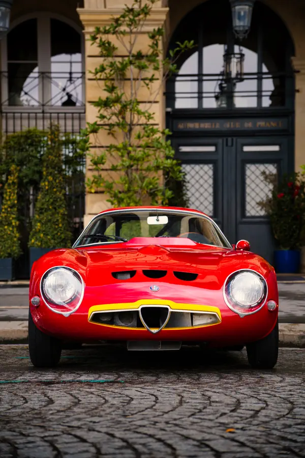 Alfa Romeo Giulia TZ rouge photographiée de face sur des pavés, avec ses phares ronds caractéristiques et sa calandre triangulaire bordée de jaune, devant l'entrée de l'Automobile Club de France.