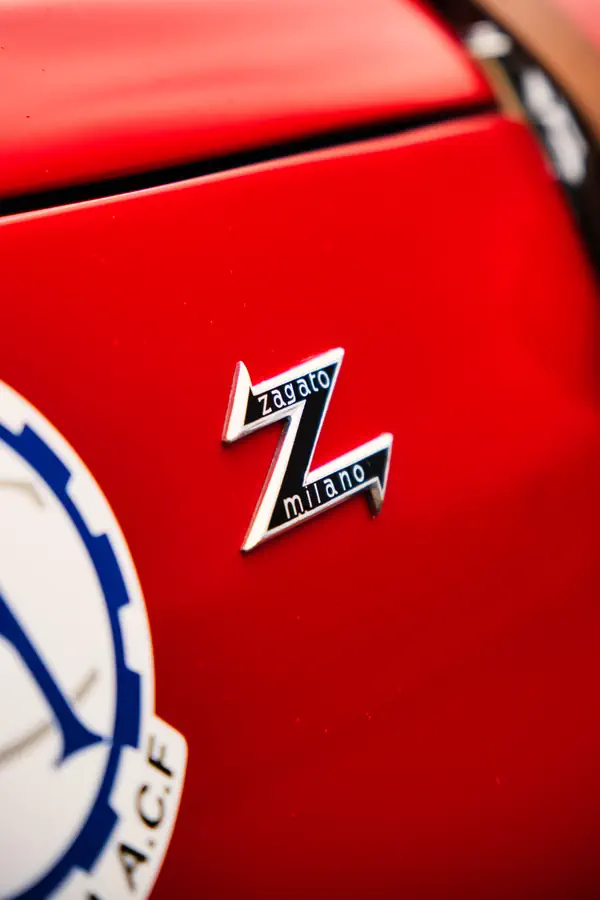 Gros plan sur l'emblème Zagato Milano en forme de Z sur la carrosserie rouge vif d'une Alfa Romeo, avec une partie du badge de l'Automobile Club de France visible à gauche.